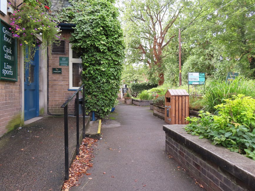 A narrow garden pathway adjacent to a brick building with a blue door, partially covered by lush green ivy climbing up the wall. The pathway, paved with asphalt, is bordered by raised flower beds fill
