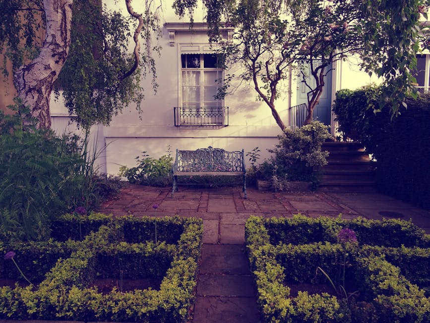 A modern communal courtyard garden adjacent to a contemporary multi-storey building with glass balconies. The garden features a variety of young trees with green and reddish leaves, planted within a r